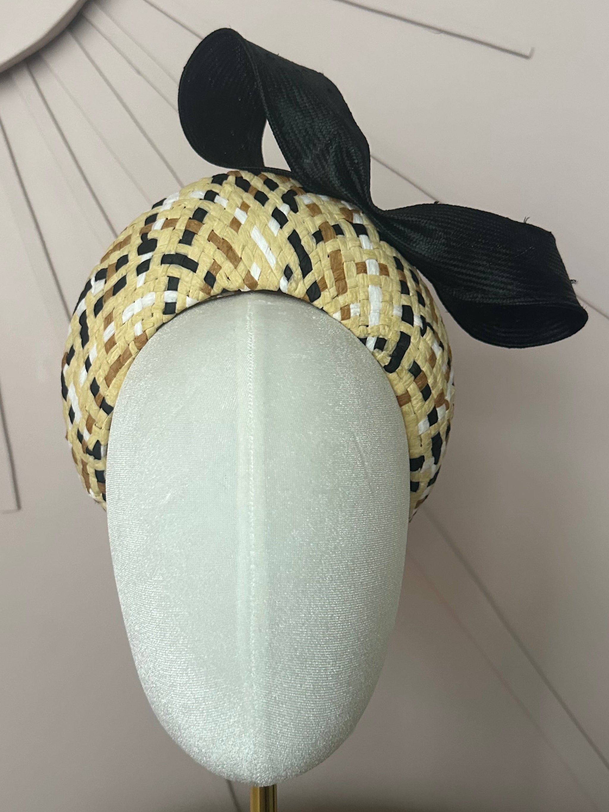 Small woven straw halo ( tall padded headband) the straw has black white and tan strands running though it. It almost creates a checkerboard effect. A black sisal bow sits at the left top side of it. The halo sits on a white velvet mannequin head in front of a pale dusky pink background with a partial sunburst that comes from the top left of the picture in an art deco style. It has a gold brassy stand with a square base on a small straw round table. 