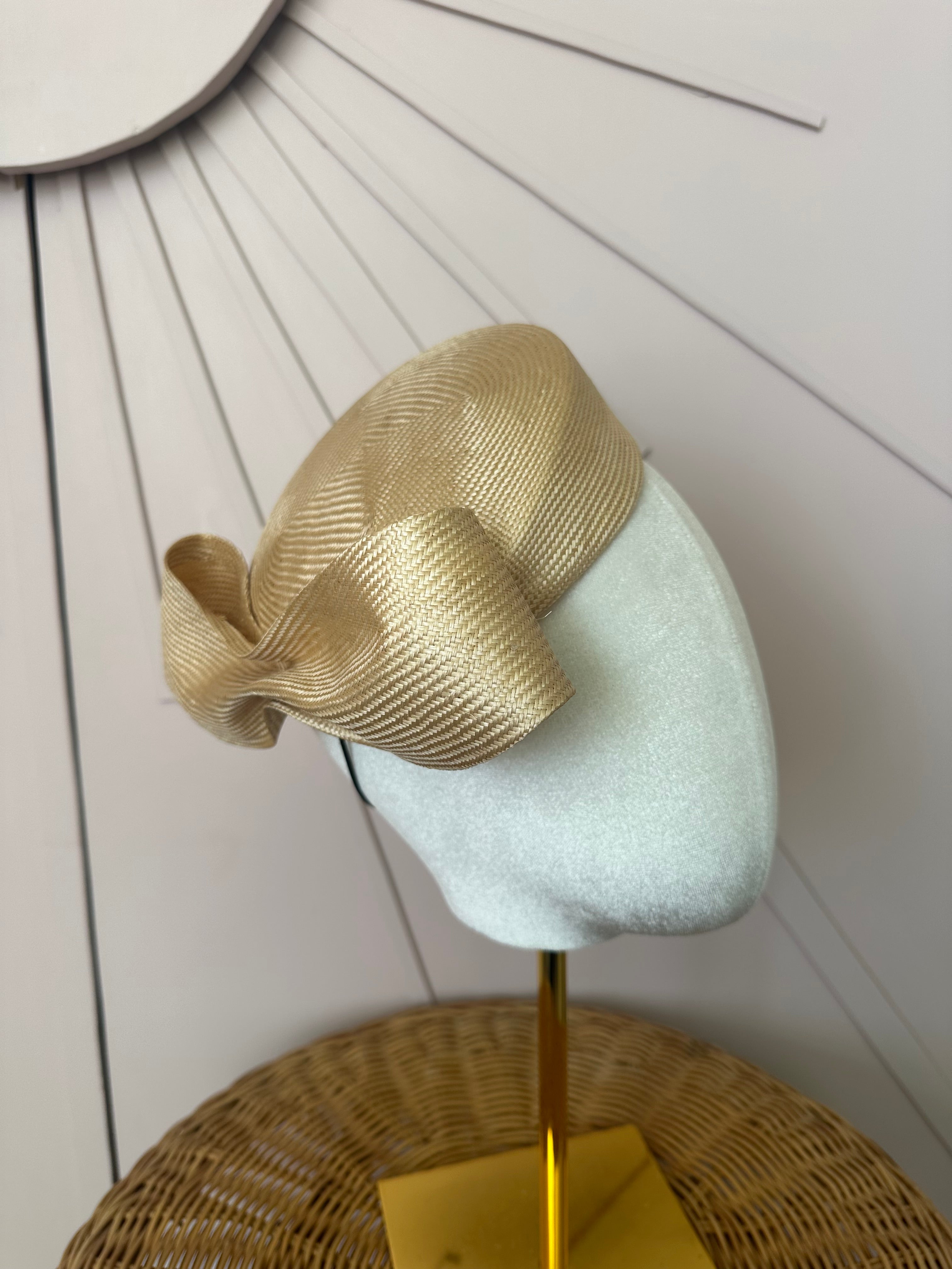 Luxury Gold sisal asymmetric pill box with a bow which sits by the ear on a white velvet mannequin head against a pale pink wooden panel that’s has an art deco star burst background. The mannequin head has a golden brassy coloured stand and sits on a cane table - very chic. 
