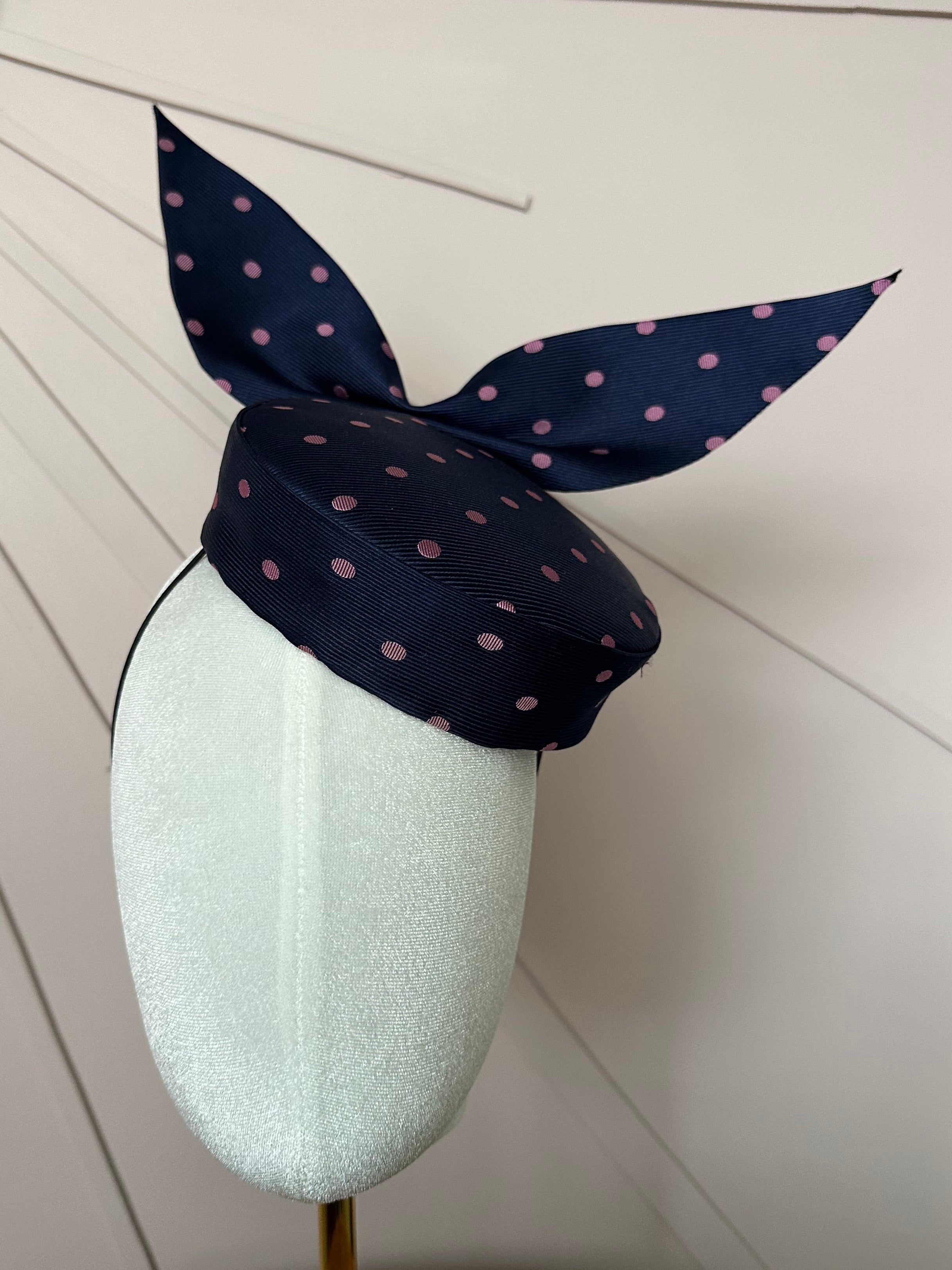 A small navy pill box with pink polka dots and a pointed bow on tip on a white velvet mannequin head with a pale dusky pink wooden background 
